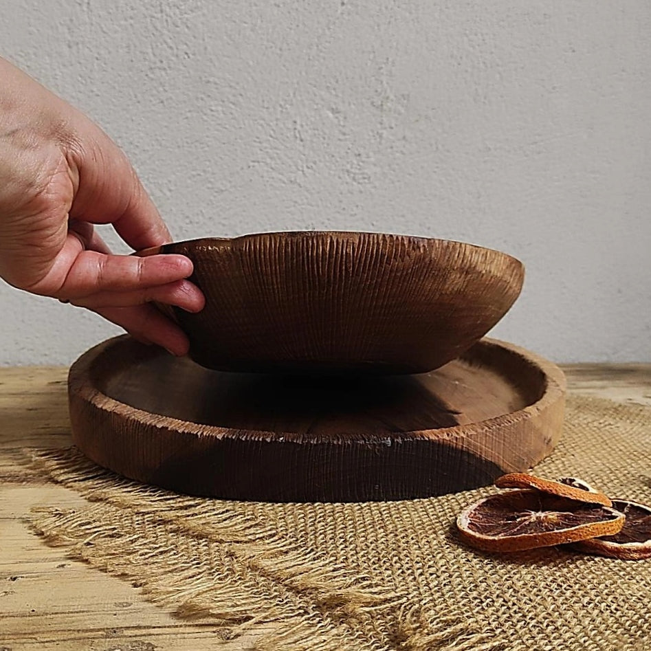 Zwei handgemachte Holzprodukte: eine gerillte Schale wird von einer Hand gehalten, darunter eine flache Holzplatte, daneben getrocknete Orangenscheiben auf Leinenstoff